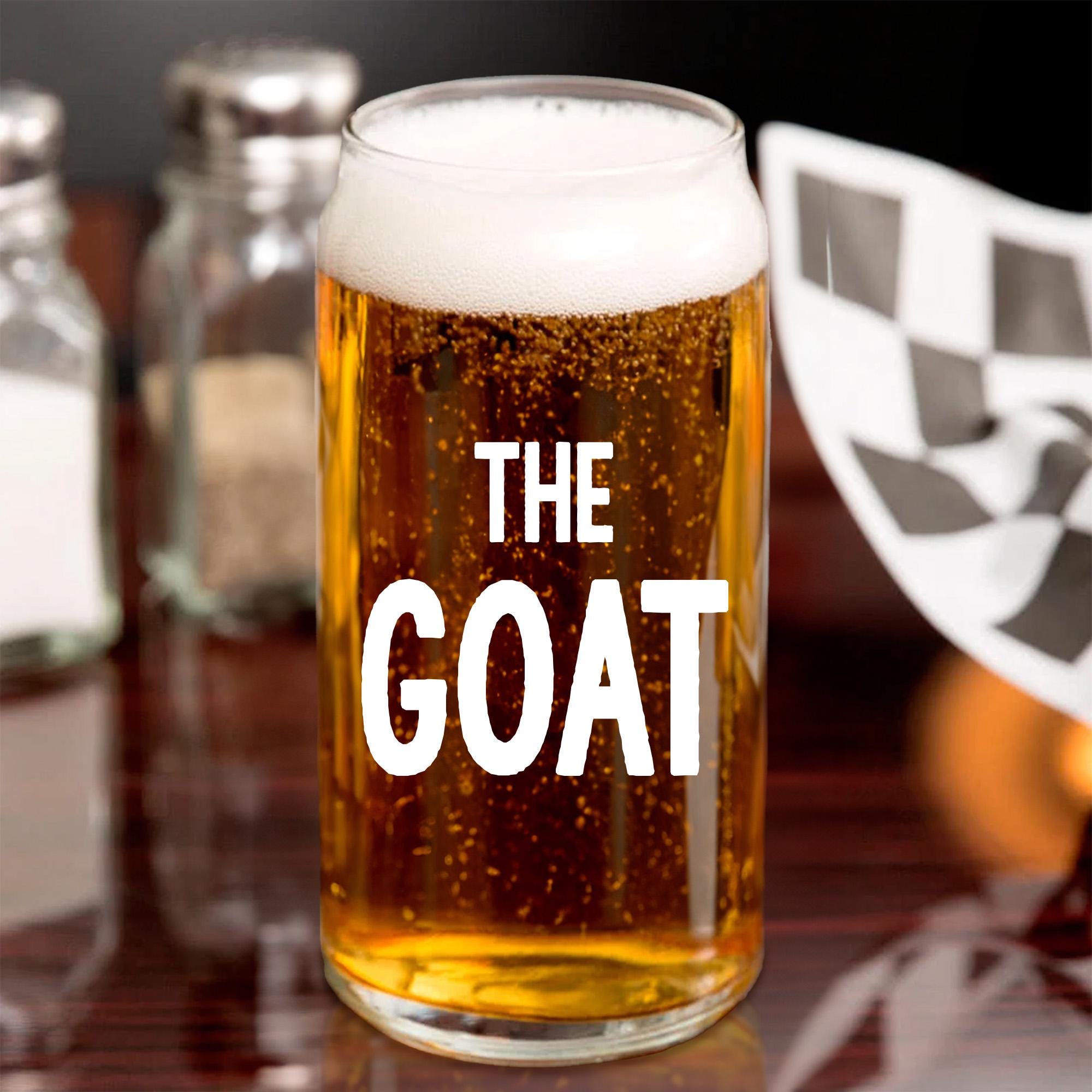 A glass of beer with foam on top, labeled &quot;THE GOAT&quot; in bold white letters, sits on a table with a checkered flag in the background.