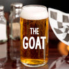 A glass of beer with foam on top, labeled "THE GOAT" in bold white letters, sits on a table with a checkered flag in the background.