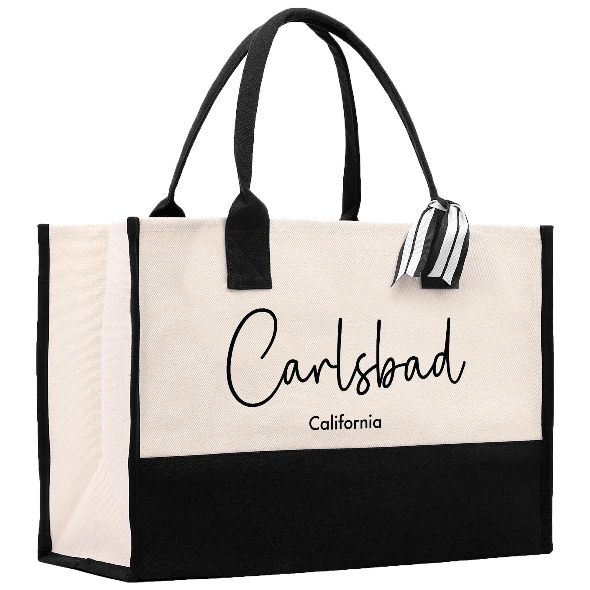 A beige tote bag with black handles and straps, featuring the text &quot;Carlsbad California&quot; printed on it.