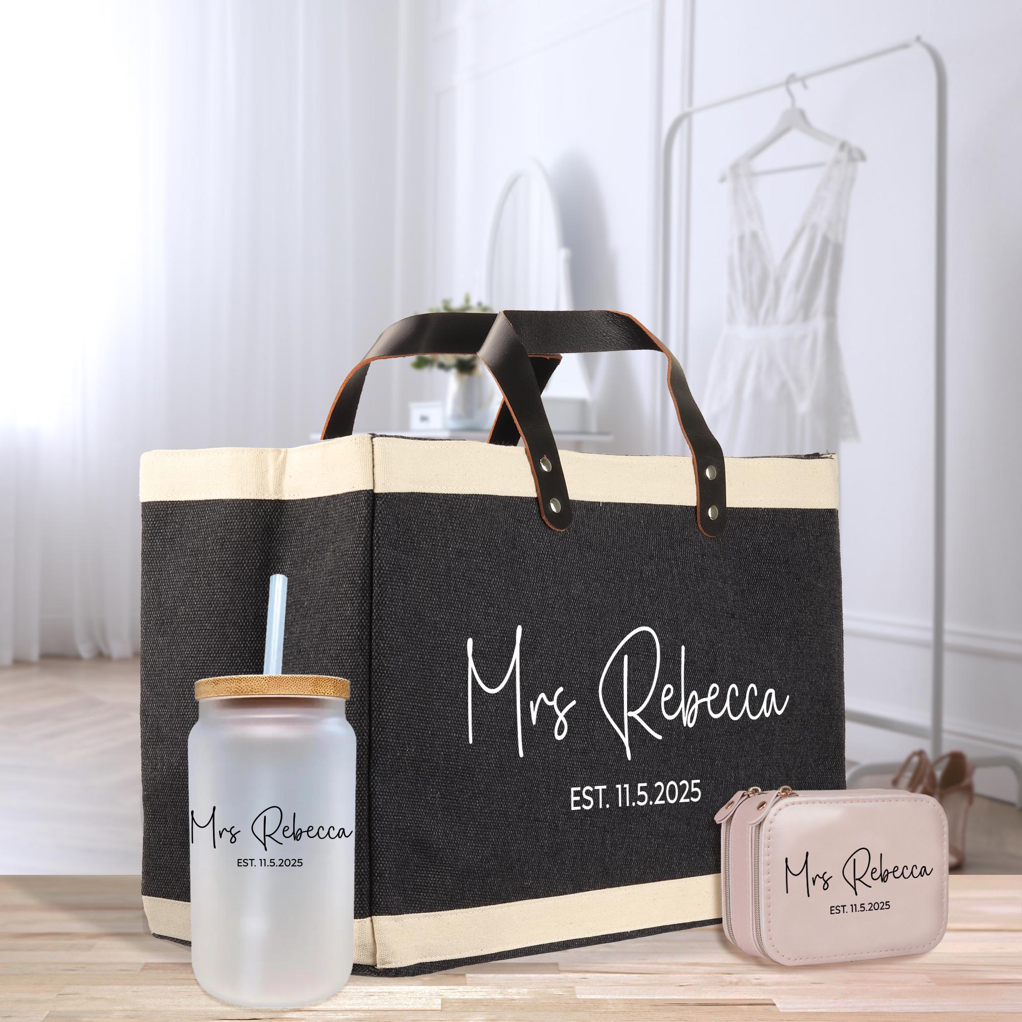 A black tote bag with a handle, a glass with a straw, and a small pink bag are displayed on a wooden surface.