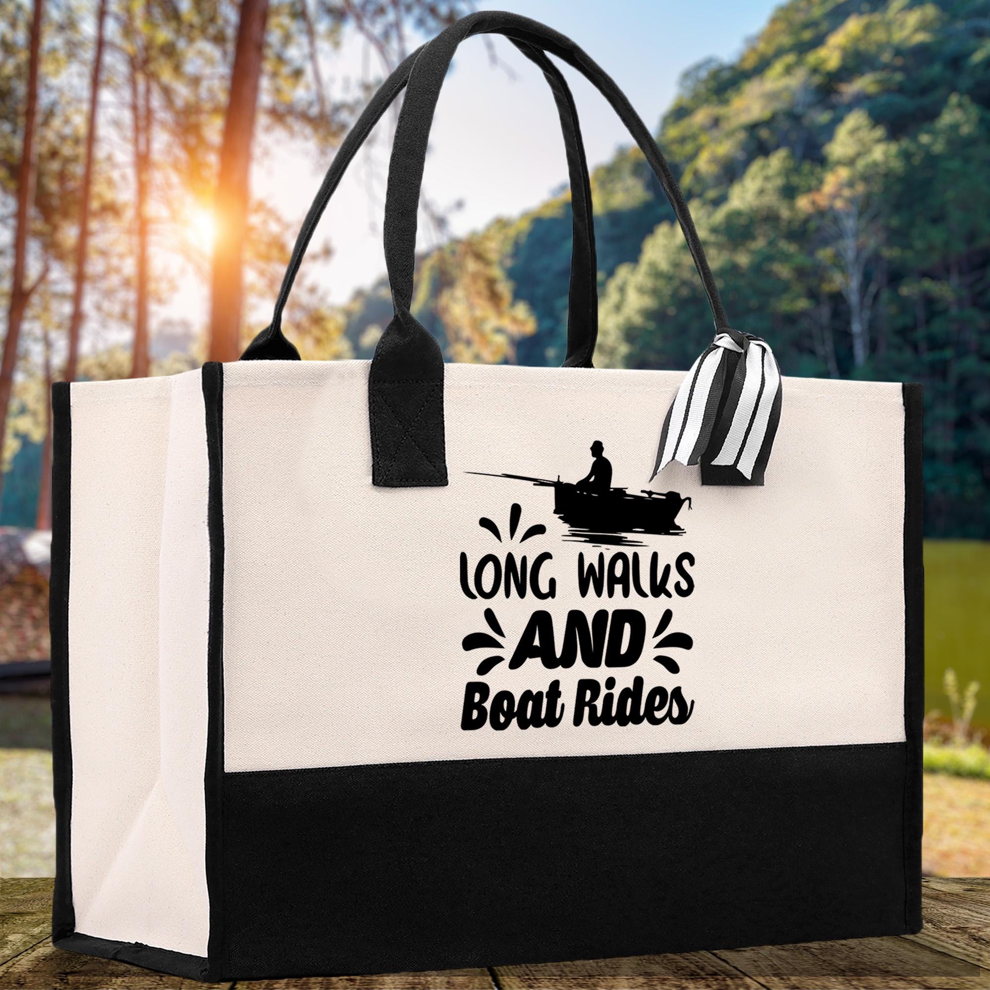 A white tote bag with a black and white graphic of a person fishing and the text "LON'G WALKS AND BOAT RIDES" printed on it, placed on a wooden surface with a natural outdoor background.