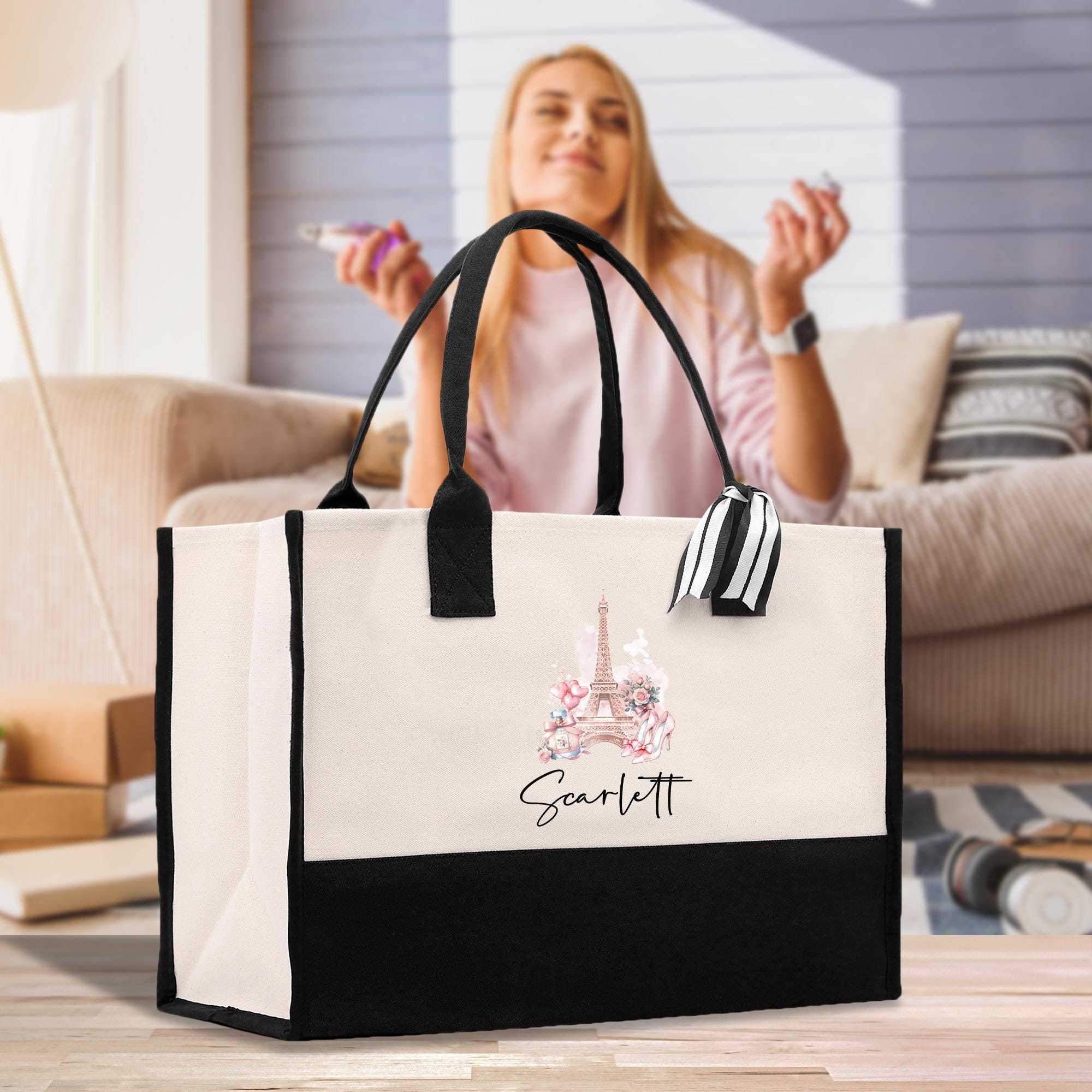 A woman is sitting on a couch in a living room, holding a black and white tote bag with a Paris-themed design.