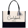 A beige tote bag with black trim and a black handle, featuring the text "Carlsbad, California" printed on it.