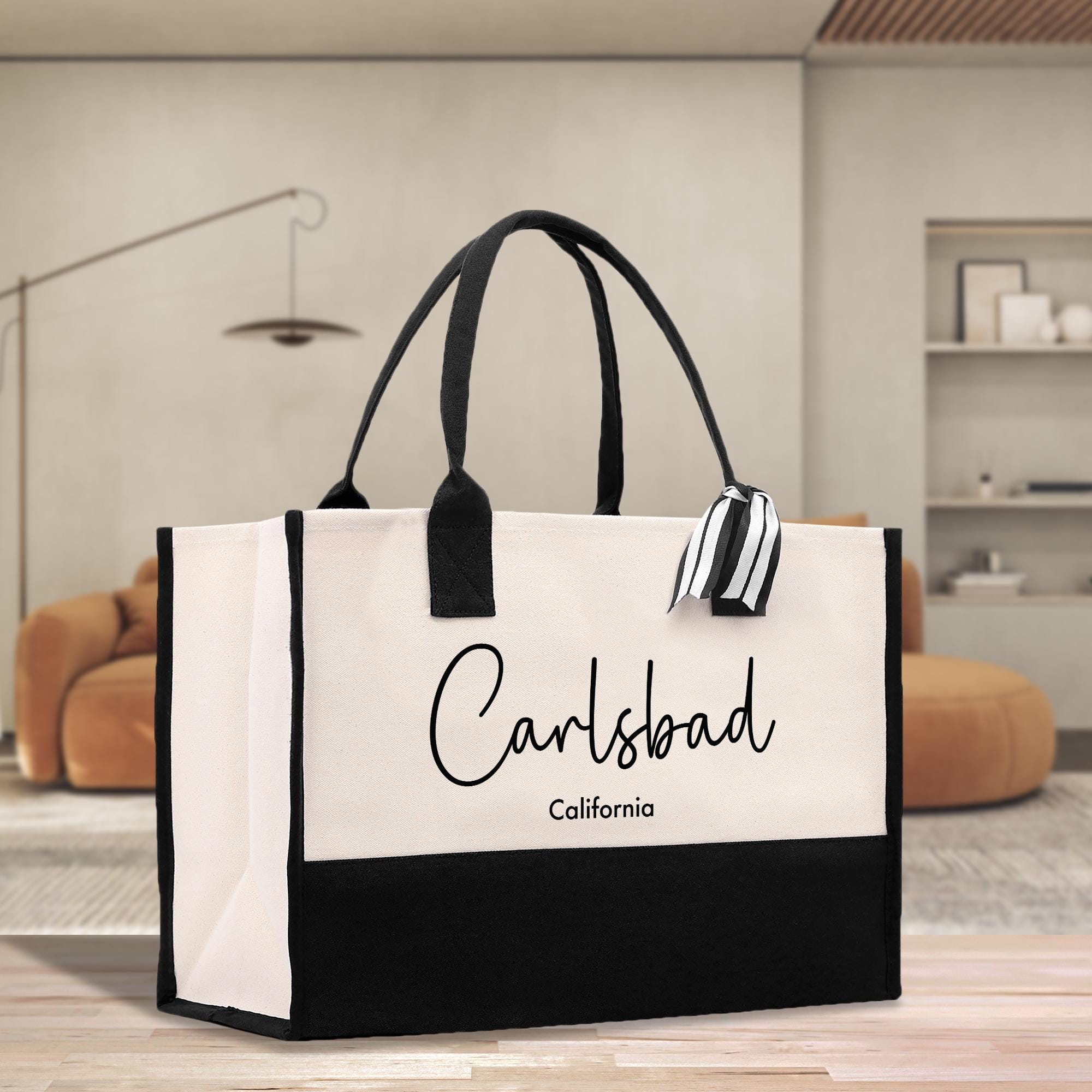 A large tote bag with the text &quot;Carlsbad California&quot; printed on it, placed on a wooden surface in a room with a brown sofa and a lamp in the background.