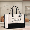 A large tote bag with the text "Carlsbad California" printed on it, placed on a wooden surface in a room with a brown sofa and a lamp in the background.
