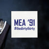 a white sign with the text "MEA '91 #the Dirty Thirty" written on it, placed on a dark surface next to a fork and knife.