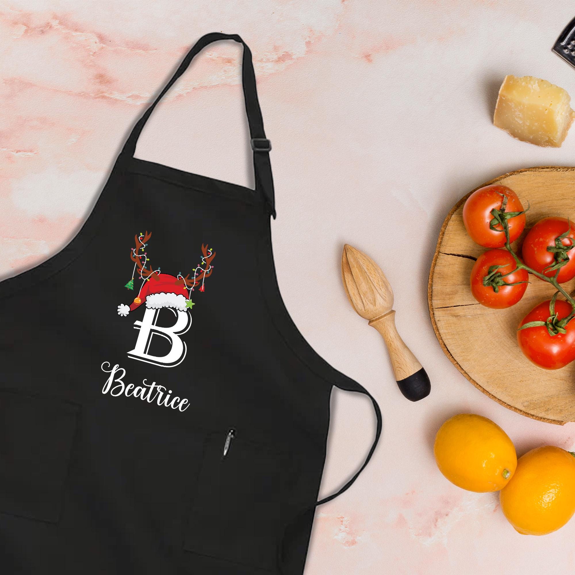 A black apron with a personalized name and a Santa hat is placed on a table next to a wooden cutting board with tomatoes, a wooden spoon, and a lemon.