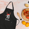 A black apron with a personalized name and a Santa hat is placed on a table next to a wooden cutting board with tomatoes, a wooden spoon, and a lemon.