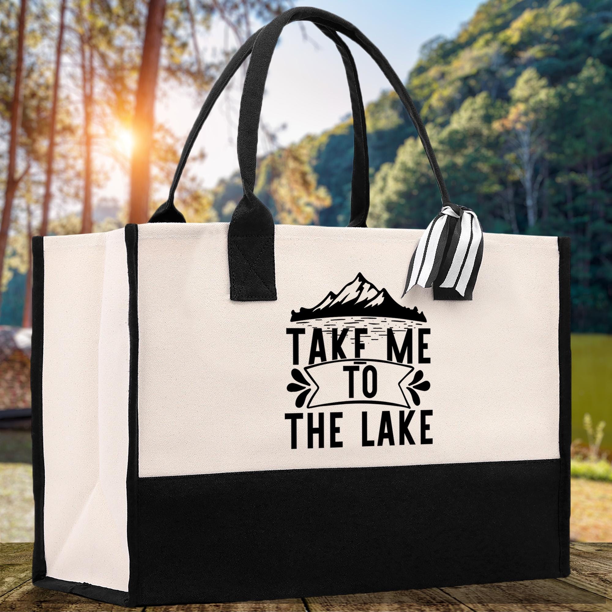 A black and white tote bag with the text "TAKE ME TO THE LAKE" printed on it, placed on a wooden surface with a natural outdoor background.