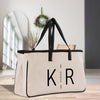 A beige tote bag with black straps and handles, featuring the initials "KR" printed on it, is placed on a wooden surface.