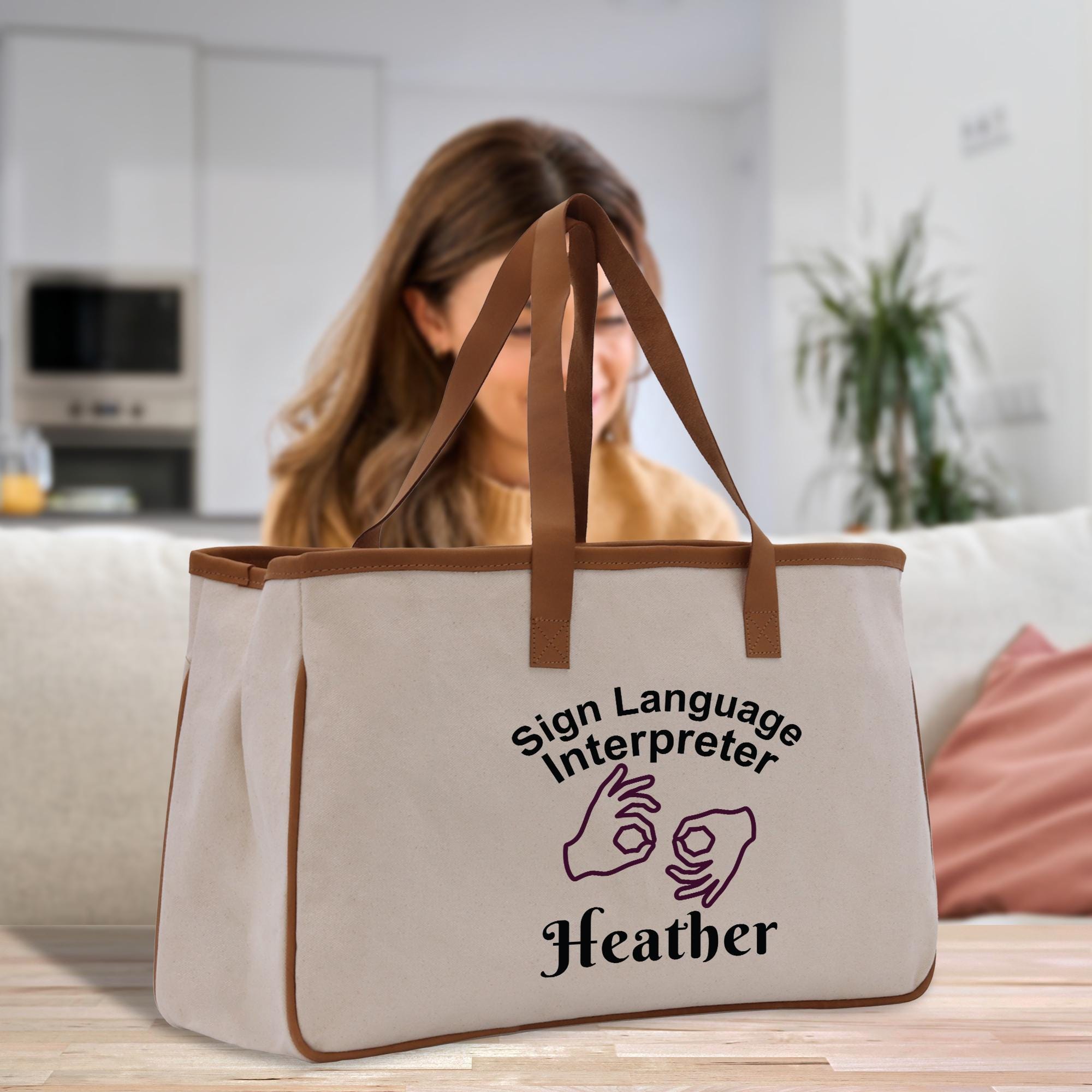 A beige tote bag with the text &quot;Sign Language Interpreter Heather&quot; printed on it, sitting on a table in a room with a woman in the background.
