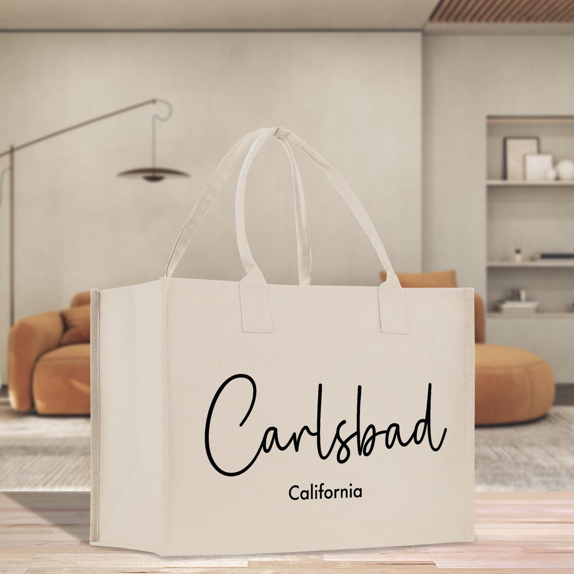 A white tote bag with the text &quot;Carlsbad California&quot; printed on it, placed on a wooden surface in a room with a brown sofa and a lamp in the background.