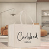 A white tote bag with the text "Carlsbad California" printed on it, placed on a wooden surface in a room with a brown sofa and a lamp in the background.