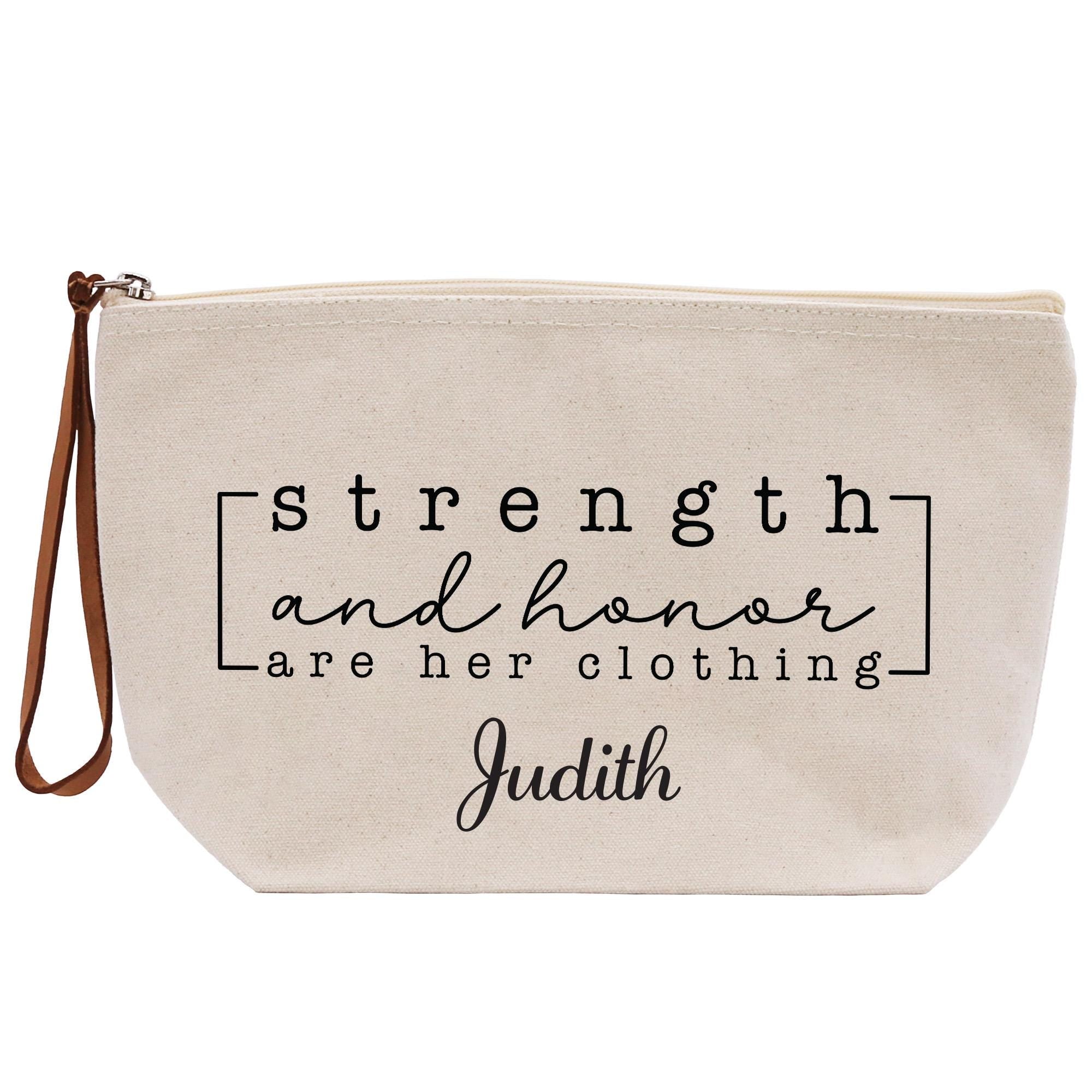 A beige canvas tote bag with the text &quot;strength and honor are her clothing&quot; printed on it.