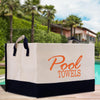 A large white and black tote bag with the text "Pool Towels" printed on it, placed on a wooden surface near a pool with palm trees in the background.