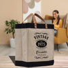 A beige tote bag with a vintage-style design, featuring text that reads "VINTAGE PREMIUM QUALITY EST. 1975 LIMITED EDITION Elizabeth's AGED 50 YEARS TO PERFECTION" and a leather handle. The bag is placed on a wooden surface, with a woman sitting in a chair in the background.
