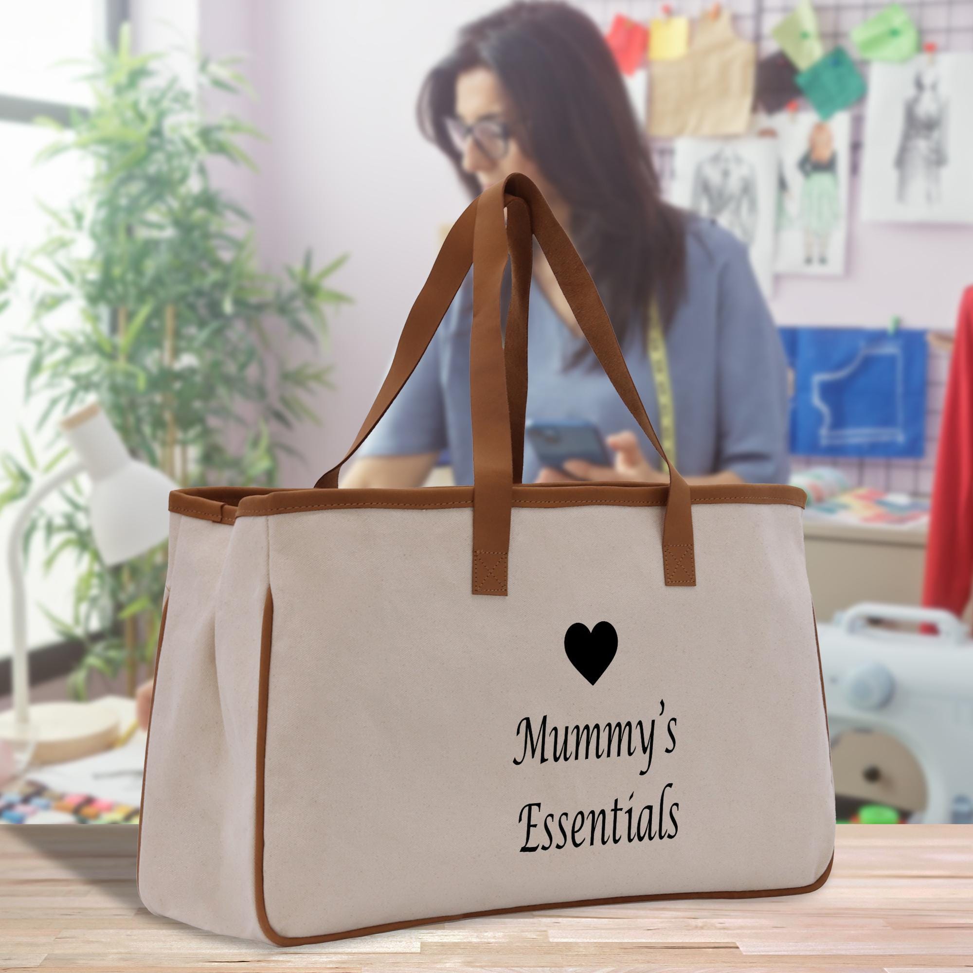 A beige canvas tote bag with the text &quot;Mummy&#39;s Essentials&quot; printed on it, placed on a wooden surface. In the background, there is a person working on a sewing machine.