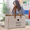 A beige canvas tote bag with the text "Mummy's Essentials" printed on it, placed on a wooden surface. In the background, there is a person working on a sewing machine.