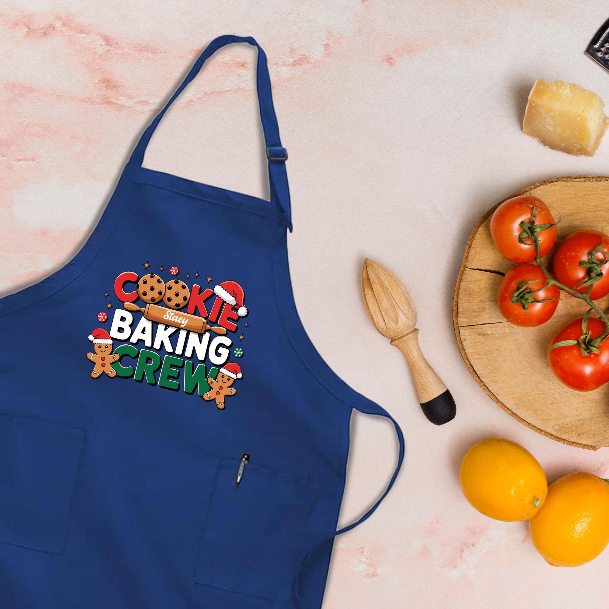 A blue apron with the text &quot;COOKIE BAKING CREW&quot; printed on it, along with various baking-related items such as a wooden whisk, a wooden cutting board with tomatoes, and a lemon.