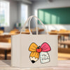 A white tote bag with a colorful bow and a pencil graphic, featuring the text "Mrs. Roberts" on it, is displayed in a blurred classroom setting.