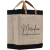A beige tote bag with black trim and leather handles, featuring a personalized text and location printed on it.