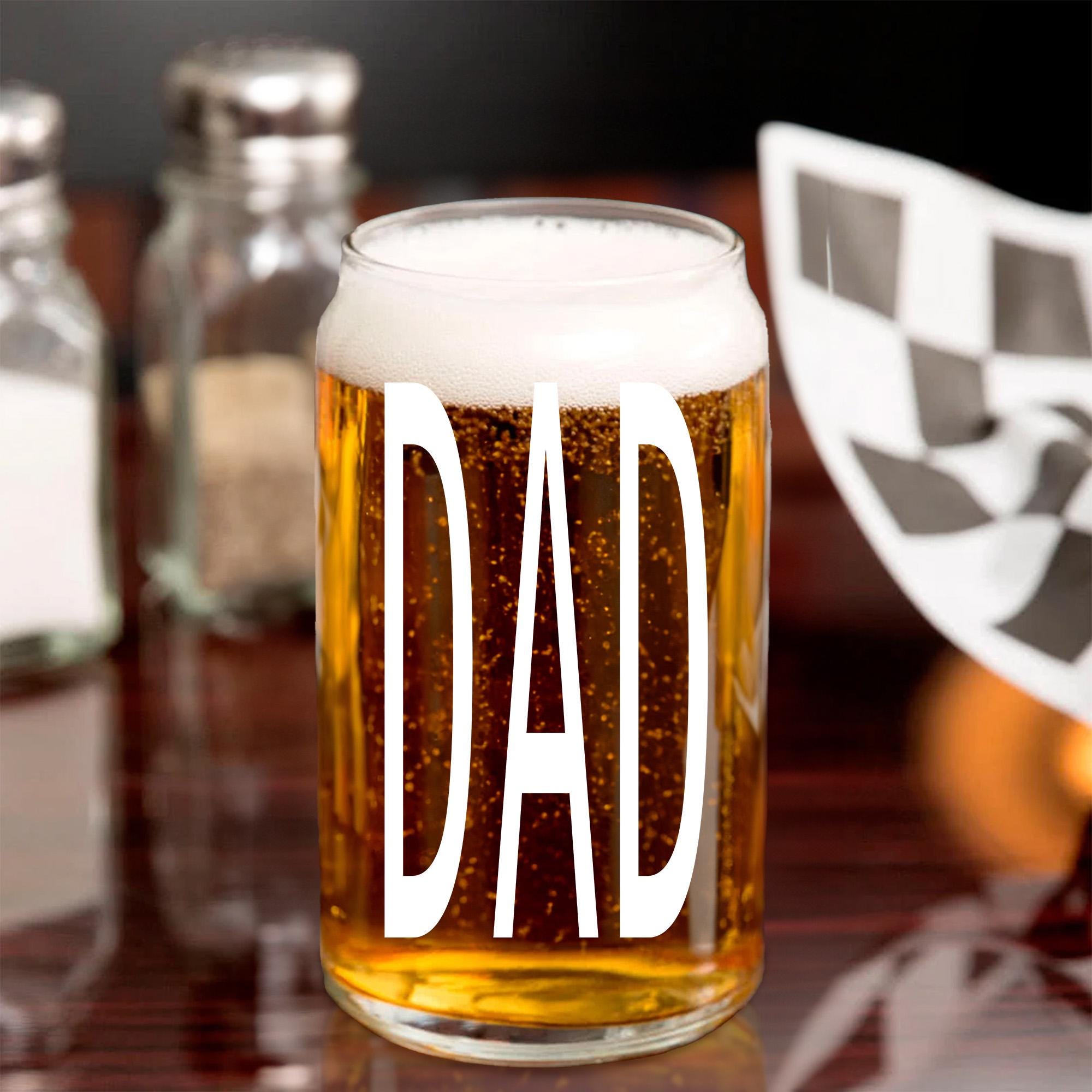 A glass of beer with the word &quot;DAD&quot; printed on it, placed on a table with other items in the background.