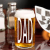 A glass of beer with the word "DAD" printed on it, placed on a table with other items in the background.