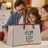 A beige canvas tote bag with a printed design on it, featuring the text "Faith Hope Love Family" and "Williams Family 2025". The bag is placed on a wooden surface, with a man and a woman standing behind it.