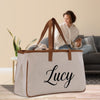 A large beige tote bag with the name "Lucy" printed on it, placed on a wooden floor in a room with a woman sitting on a couch in the background.