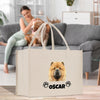 A woman is sitting on a couch in a living room, petting a dog. A large white tote bag with a dog image and the name "Oscar" printed on it is placed in front of the couch.