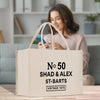 A beige tote bag with text printed on it, placed on a wooden surface. In the background, a woman is sitting on a couch and looking at her phone.