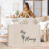 A beige tote bag with a floral design and the word "Manny" printed on it, placed on a wooden surface in a living room setting.