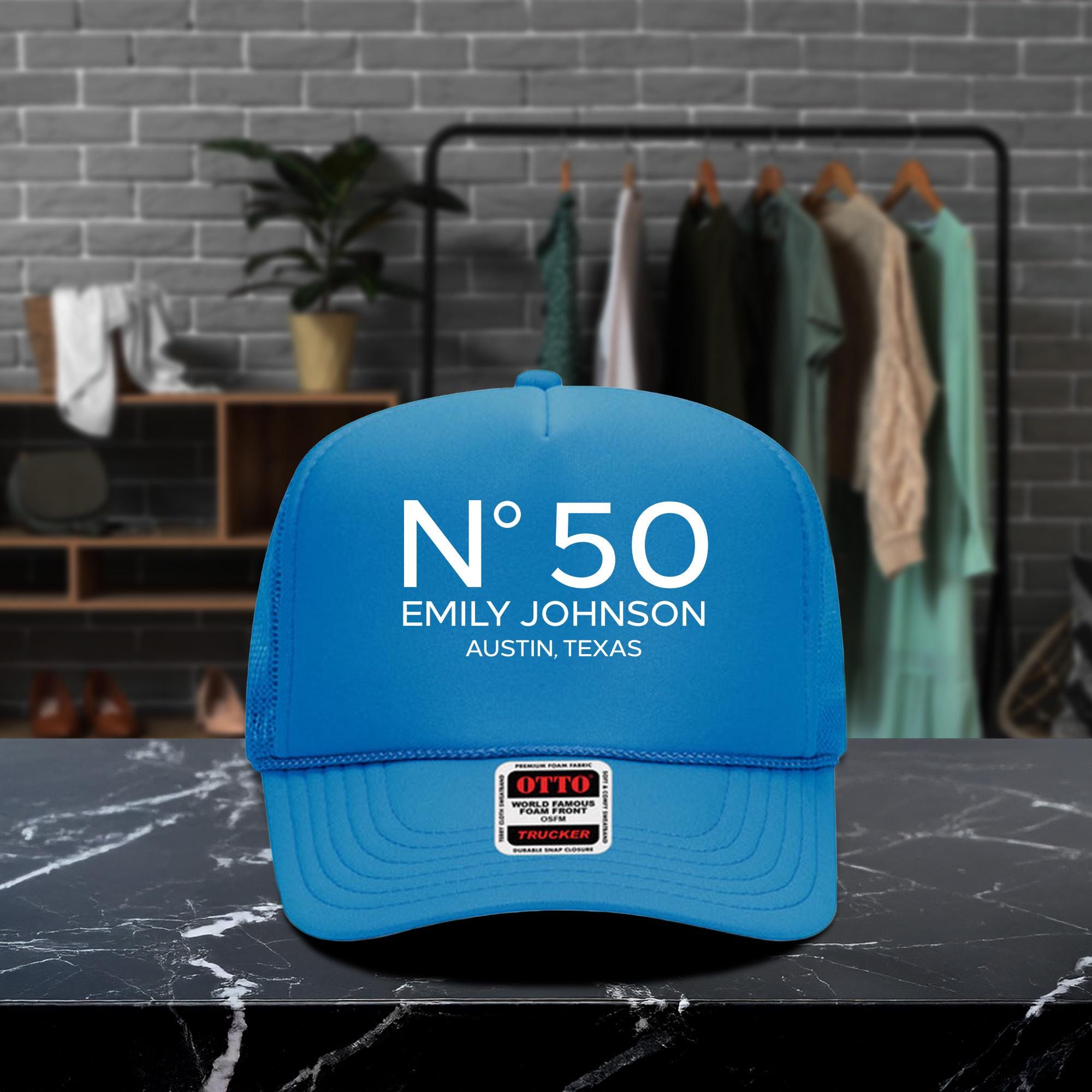 A blue baseball cap with the text &quot;N° 50 EMILY JOHNSON AUSTIN, TEXAS&quot; printed on it, placed on a marble countertop.