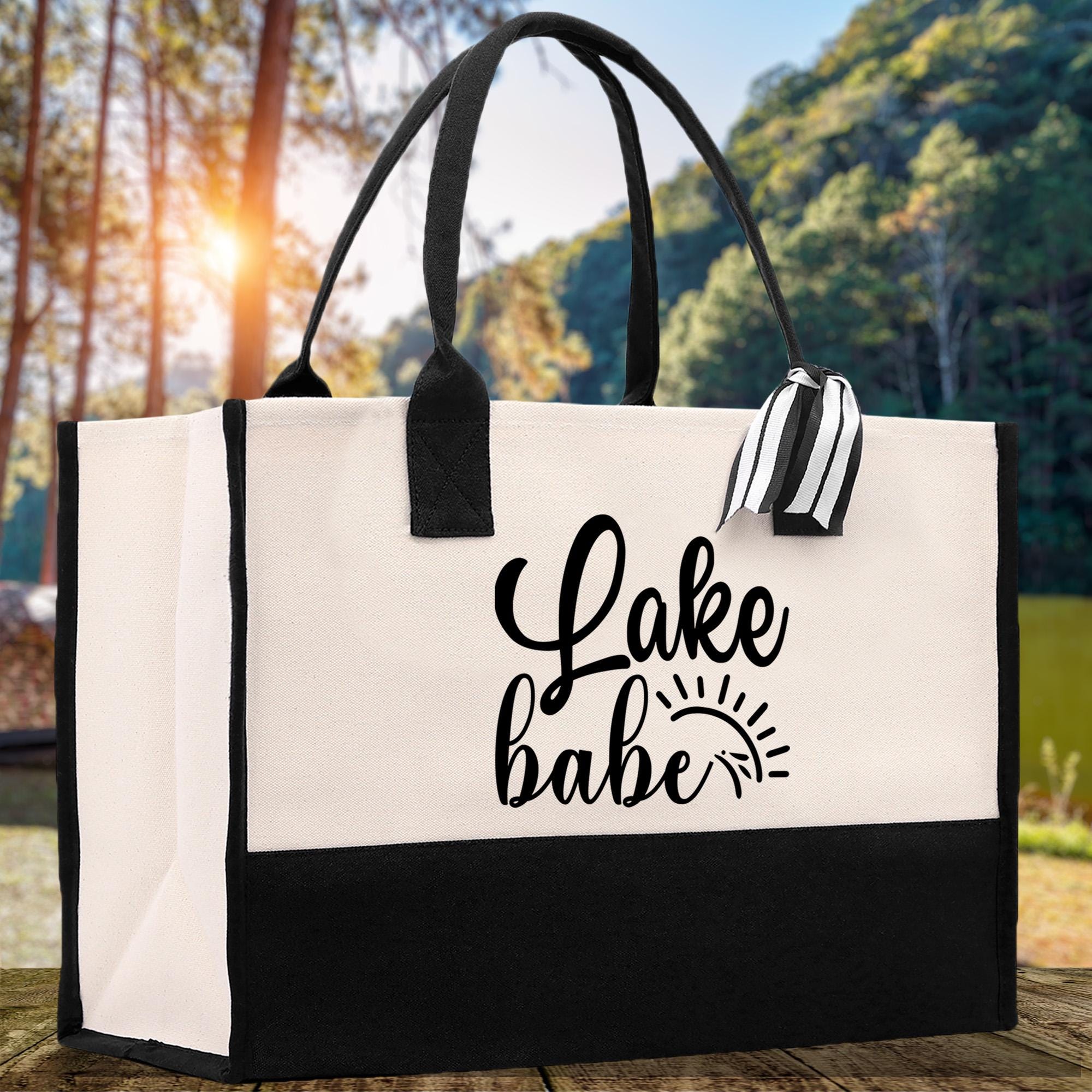 A black and white tote bag with the text "Lake Babe" printed on it, placed on a wooden surface with a natural outdoor background.