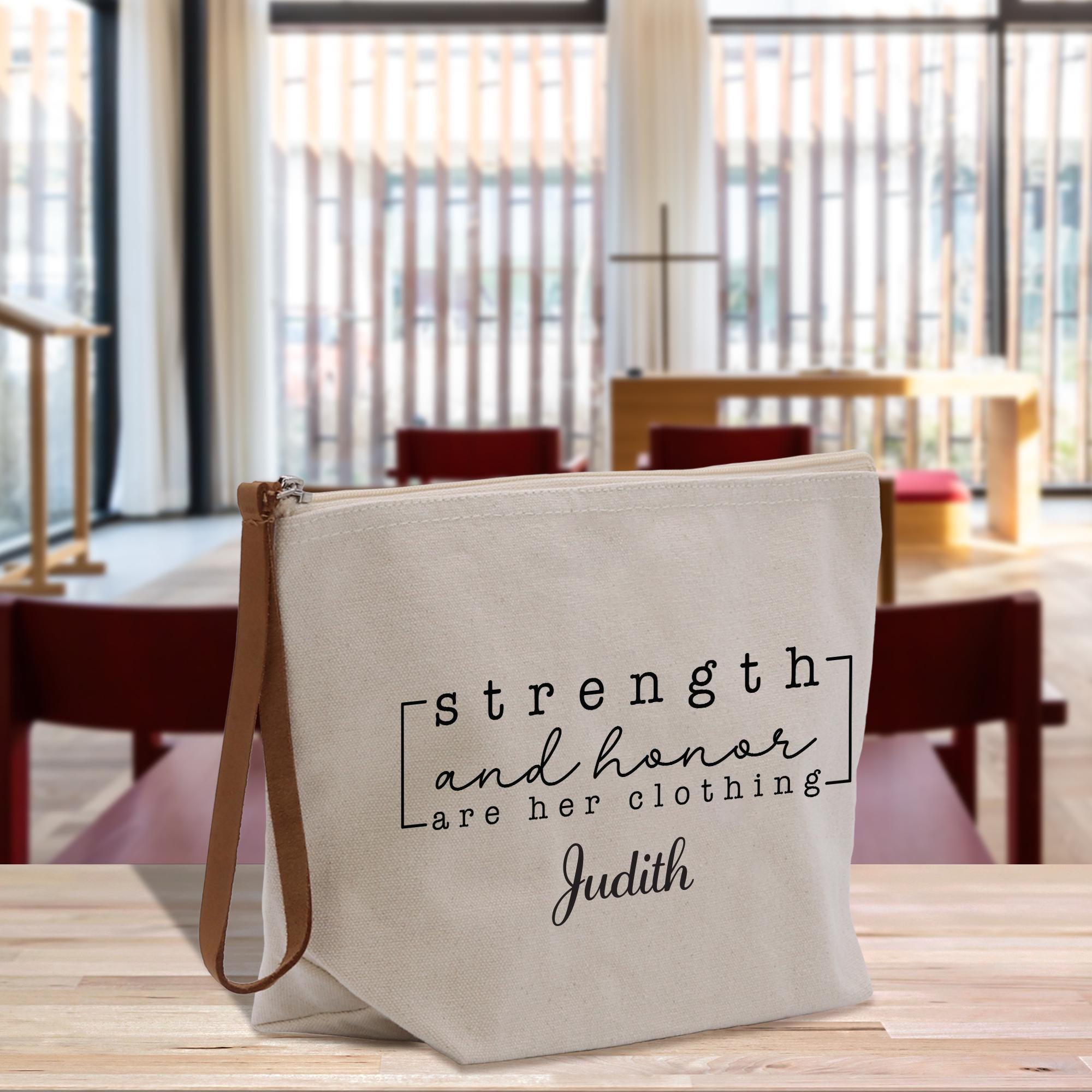 A beige tote bag with the text &quot;strength and honor are her clothing&quot; printed on it, along with the name &quot;Judith&quot; below.