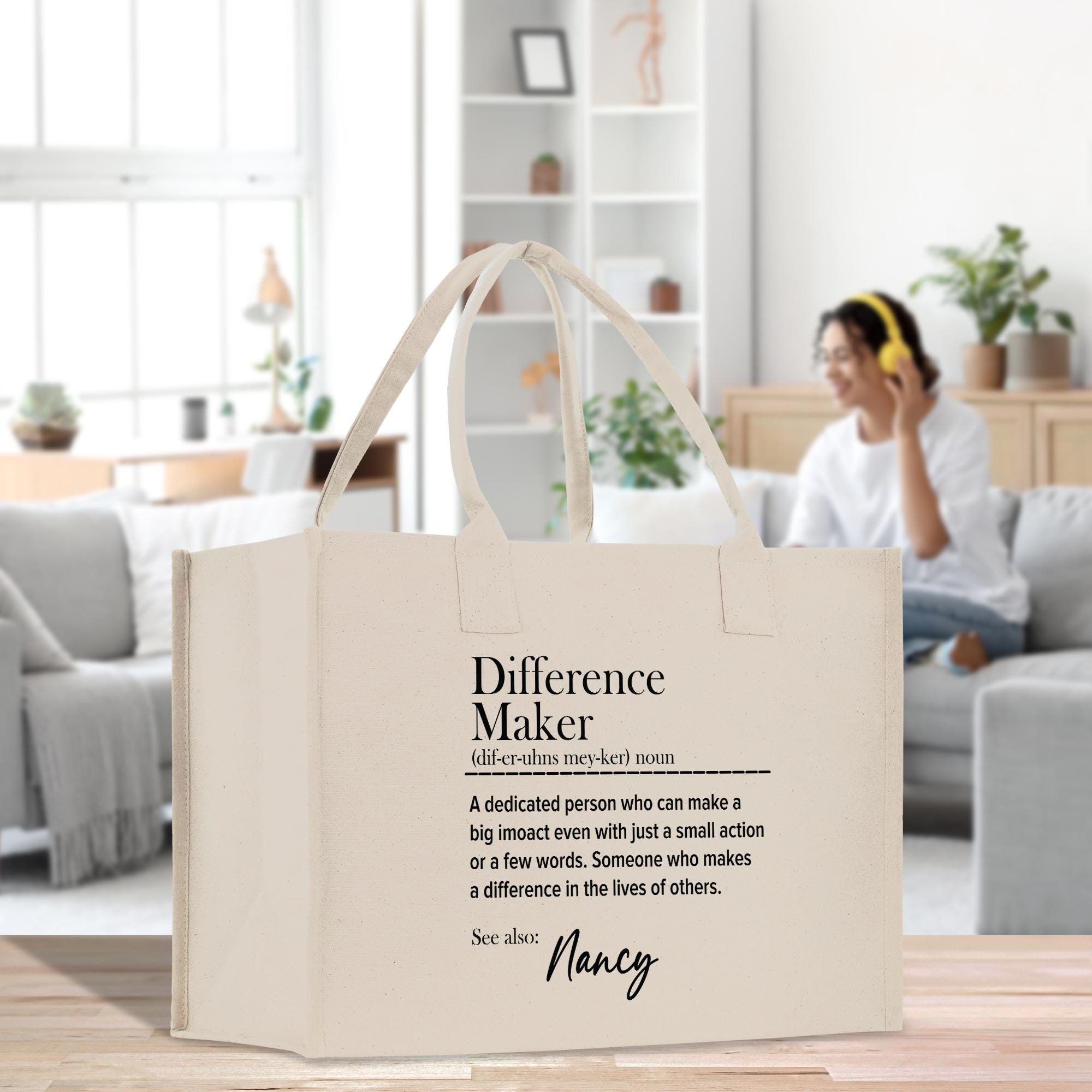 a beige tote bag with text printed on it, placed on a wooden surface in a room with a couch and potted plants in the background.