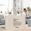 a beige tote bag with text printed on it, placed on a wooden surface in a room with a couch and potted plants in the background.