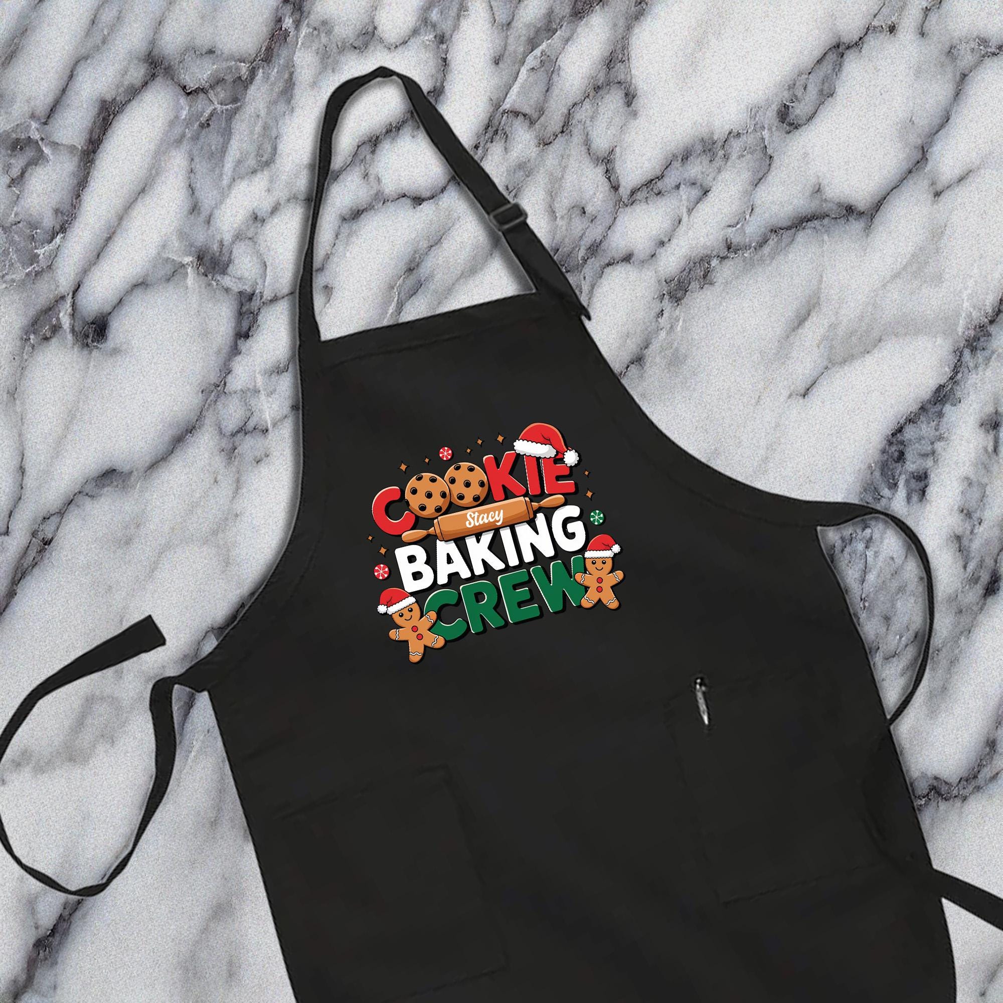A black apron with a festive design, featuring the text &quot;COOKIE BAKING CREW&quot; and images of cookies and gingerbread men.