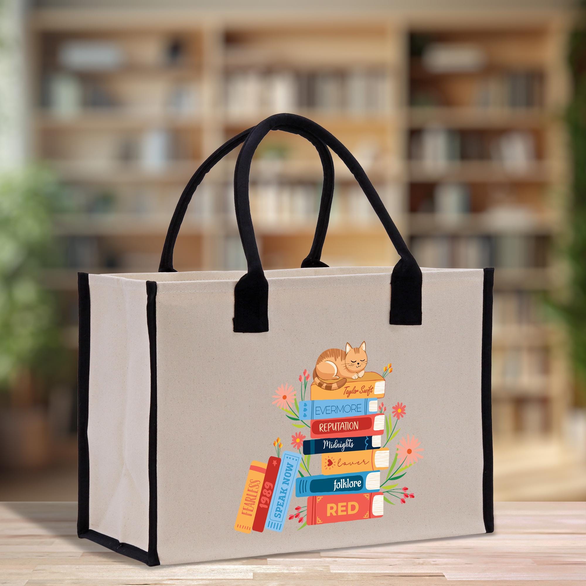 A beige tote bag with a printed design of a stack of colorful books and a cat on top, placed on a wooden surface.