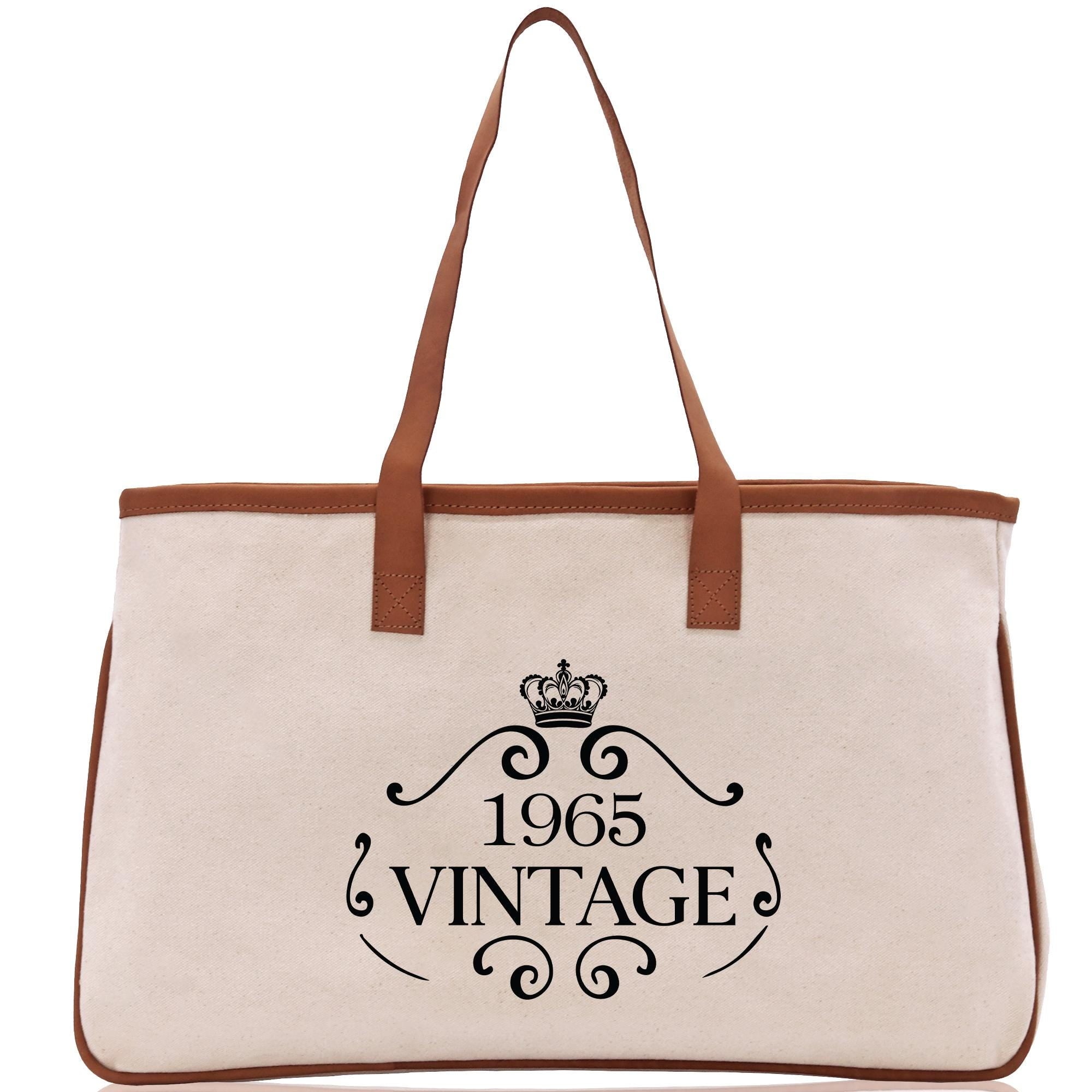 A beige canvas tote bag with a brown leather strap and a black text design that reads "1965 VINTAGE".