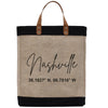 A beige tote bag with black trim and leather handles, featuring the text "Nashville" and a coordinate pair "36.1627 N, 86.7816 W" printed on it.