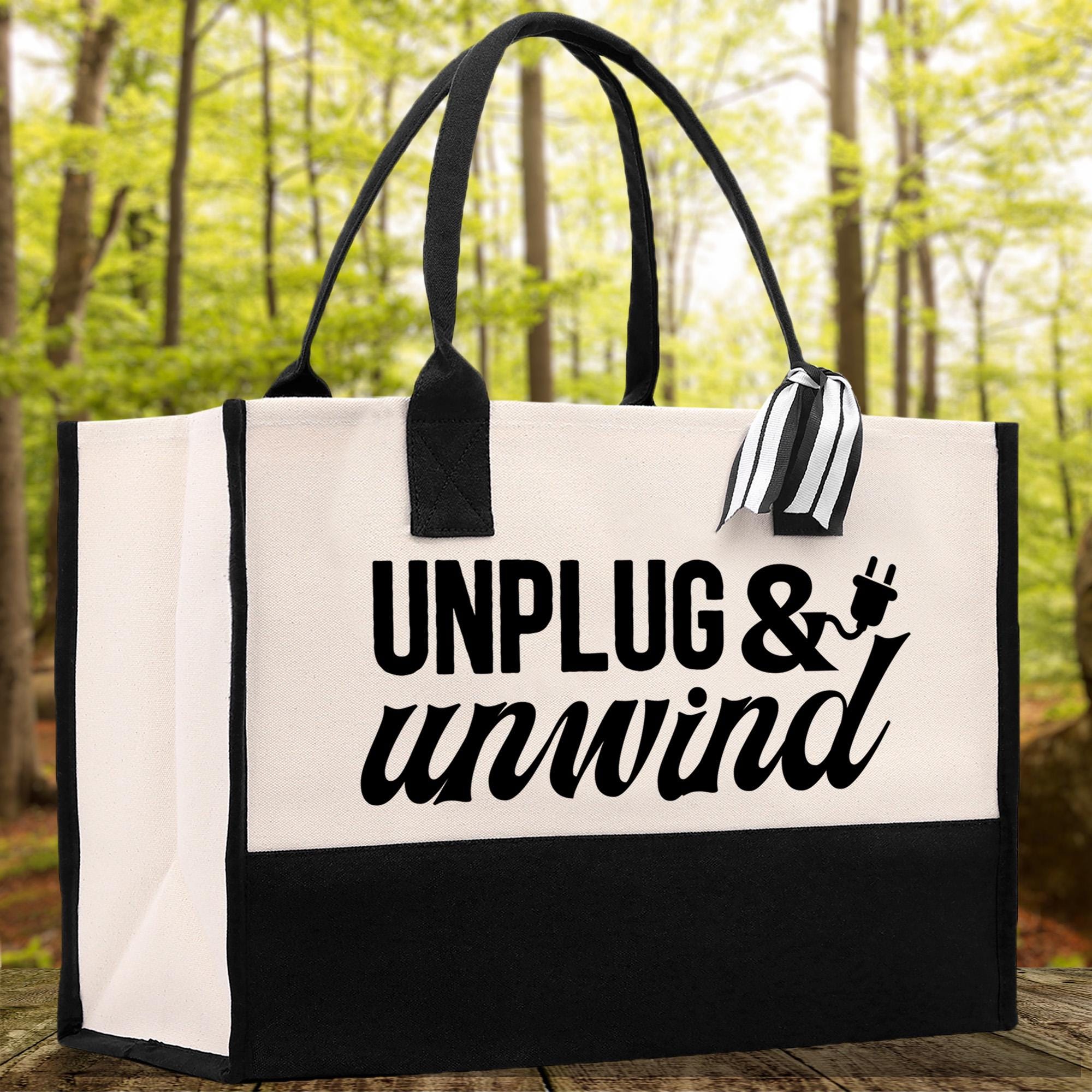 A black and white tote bag with the text "UNPLUG & unwind" printed on it, placed on a wooden surface against a blurred background of trees.