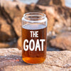 A glass jar filled with a brown liquid, with the word "THE GOAT" printed on it, placed on a rock.