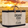 A beige canvas tote bag with black straps and handles, featuring the text "ORGANIC FOOD just kicking IT'S WINE" printed on it, placed on a wooden surface against a backdrop of a sunset over a field.