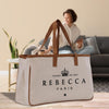 a large beige tote bag with the text "REBECCA PARIS" printed on it, placed on a wooden surface. In the background, there is a woman sitting on a couch, and a potted plant can be seen nearby.