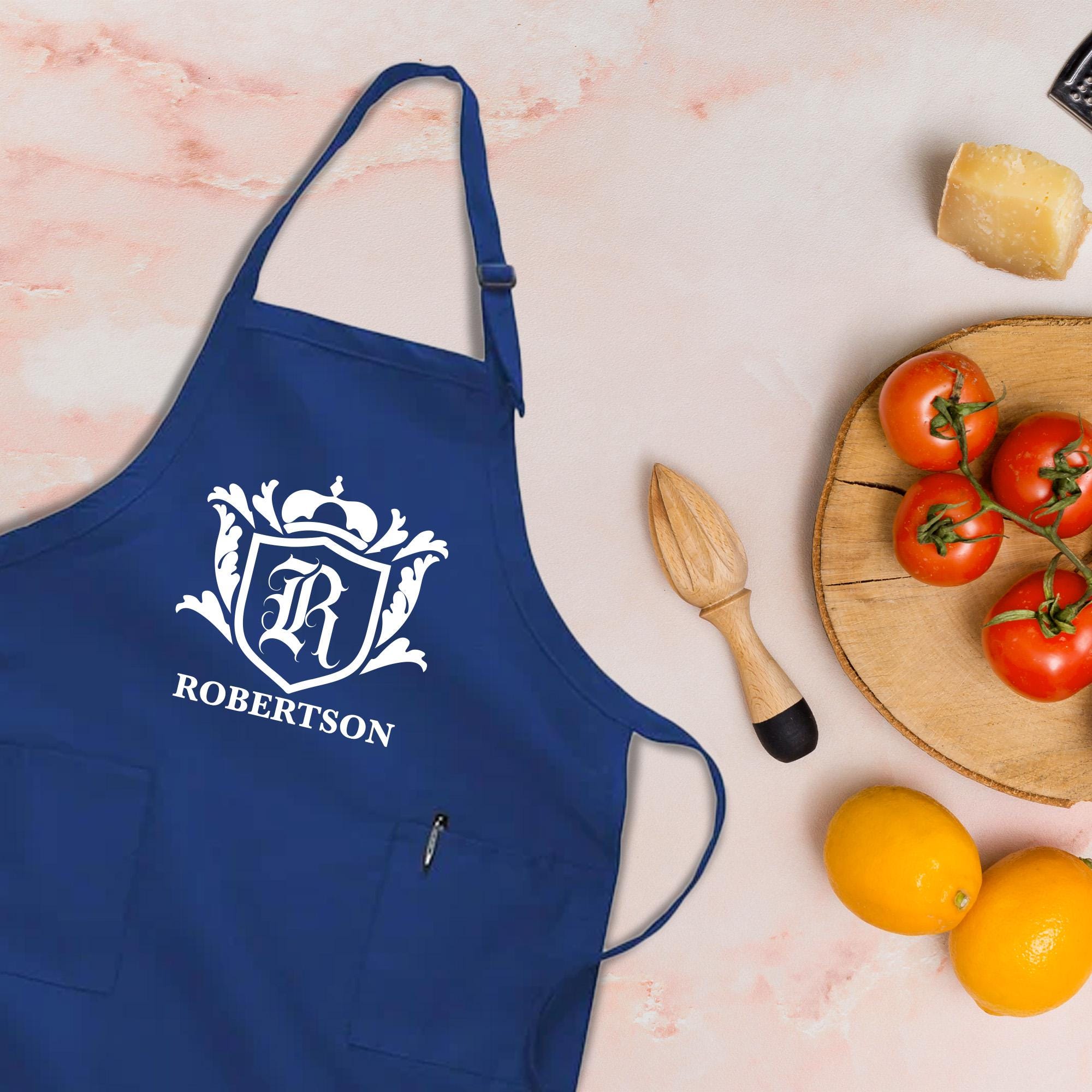 A blue apron with a white monogrammed &quot;R&quot; logo, a wooden citrus juicer, and a plate of fresh tomatoes and oranges on a marble surface.