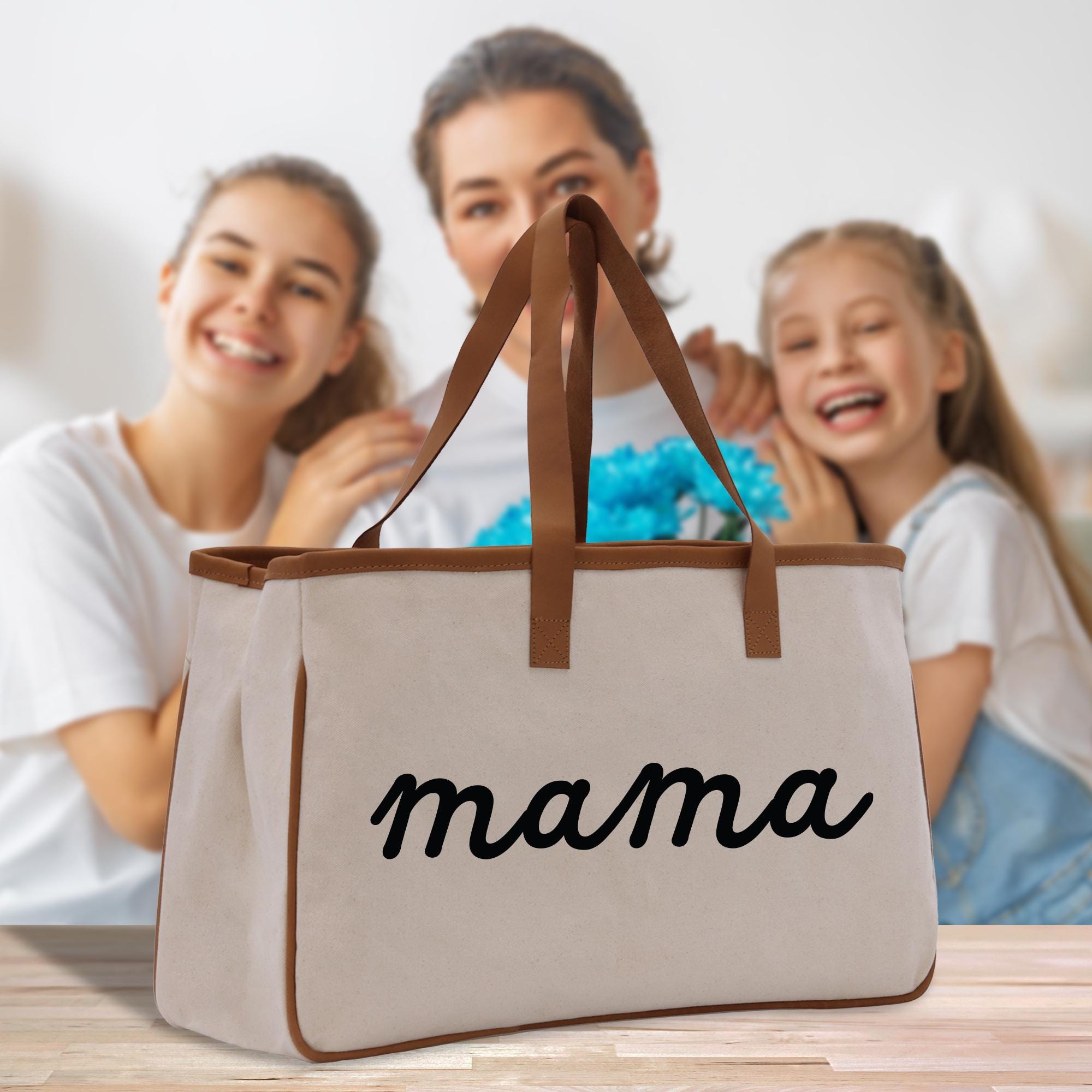 A beige tote bag with the word &quot;mama&quot; printed on it, placed on a wooden surface. In the background, there are two smiling women, one of whom is holding a blue flower.