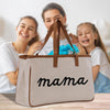 A beige tote bag with the word "mama" printed on it, placed on a wooden surface. In the background, there are two smiling women, one of whom is holding a blue flower.