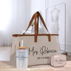 A beige tote bag with the name "Mrs Rebecca" printed on it, along with a straw cup and a small purse, all placed on a wooden surface.