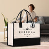 A large white tote bag with black trim and the text "REBECCA PARIS" printed on it, placed on a wooden floor in a room with a woman sitting on a couch in the background.